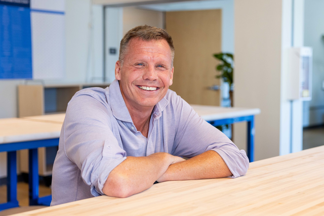 A smiling middle-aged man with short hair sits at a light wooden table in a modern, bright room, wearing a light purple button-up shirt.